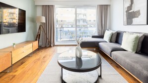 A modern living room with a sofa, a coffee table, a TV, and a large window with a view of buildings. - Luxurious Apartment in Wandsworth (London)