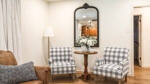 Un salon avec un canapé en cuir marron, deux fauteuils à motifs écossais, une table ronde en bois ornée d’un vase de fleurs et un grand miroir accroché au mur.