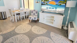 Et værelse med spiseplads, et fjernsyn der viser 'OCMD GETAWAYS', en stribet måtte og strandinspireret vægkunst.