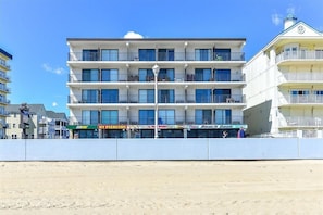 Un hôtel à plusieurs étages doté de balcons, offrant une vue sur la plage et abritant divers commerces, notamment un restaurant Subway et un salon de piercing.