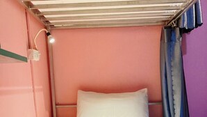 A single bed with white linens, a heart-shaped towel arrangement, a wall-mounted light, and a window with blue curtains. - Luang Prabang Hostel (Luang Prabang)