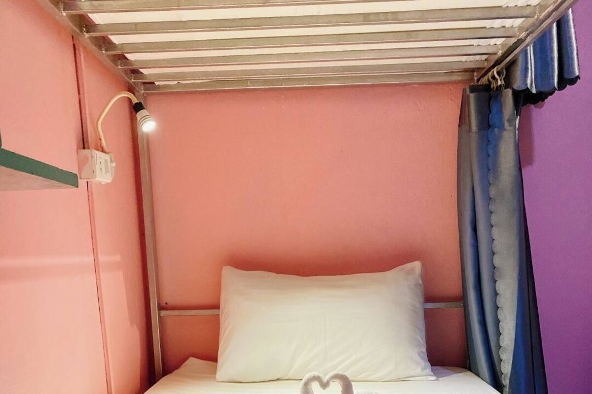 A single bed with white linens, a heart-shaped towel arrangement, a wall-mounted light, and a window with blue curtains.