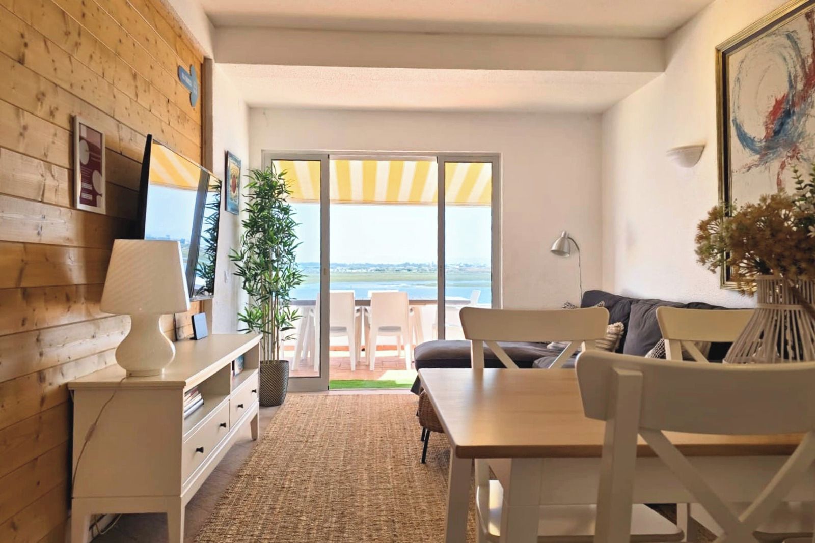 A room with a wooden wall, a desk with a lamp, a chair, a sofa, and a view through a glass door to the outside.