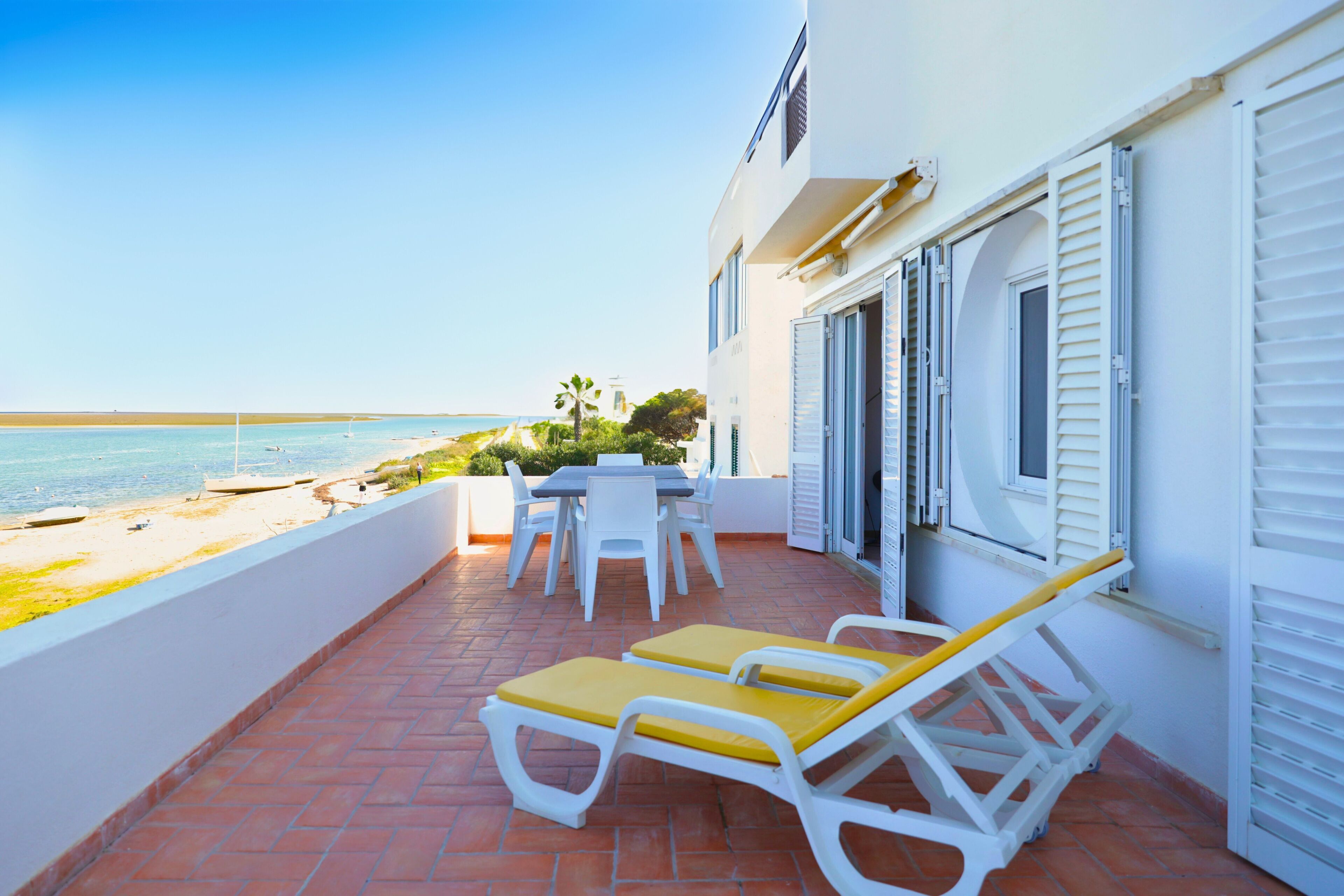 A terrace with a table and chairs, a pair of sun loungers, and a view of the beach and ocean.