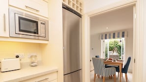 A kitchen with a stainless steel refrigerator, a microwave, and a Bosch toaster. There is a dining area with a table and chairs visible through an open door. - November Cottage (Dartmouth)
