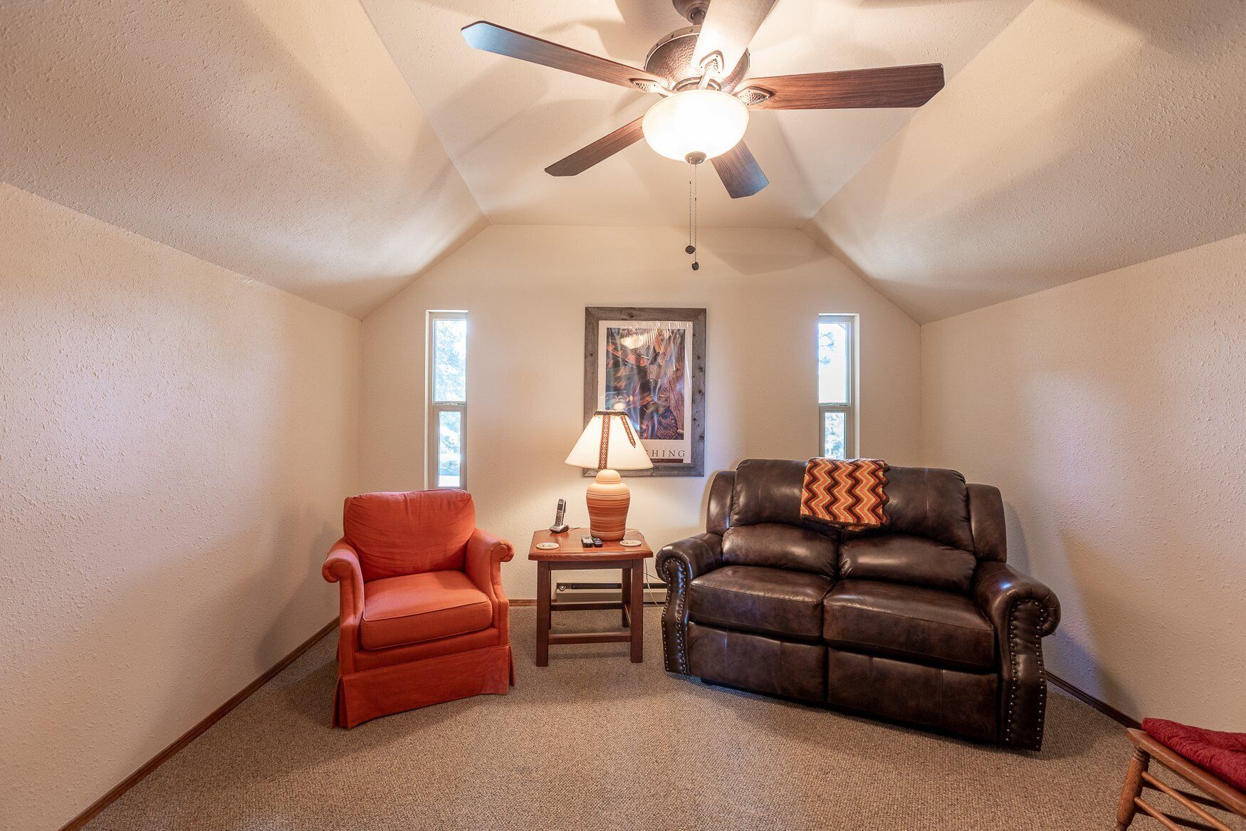 A cozy attic room with a sloped ceiling, two windows, a ceiling fan, a red armchair, a brown leather couch, and a small table with a lamp.