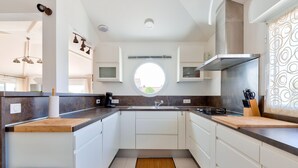 A modern kitchen with white cabinets, a central island, and a round window above the sink. - Le Fenouiller, Spacious house for 6, swimming pool (Le Fenouiller)