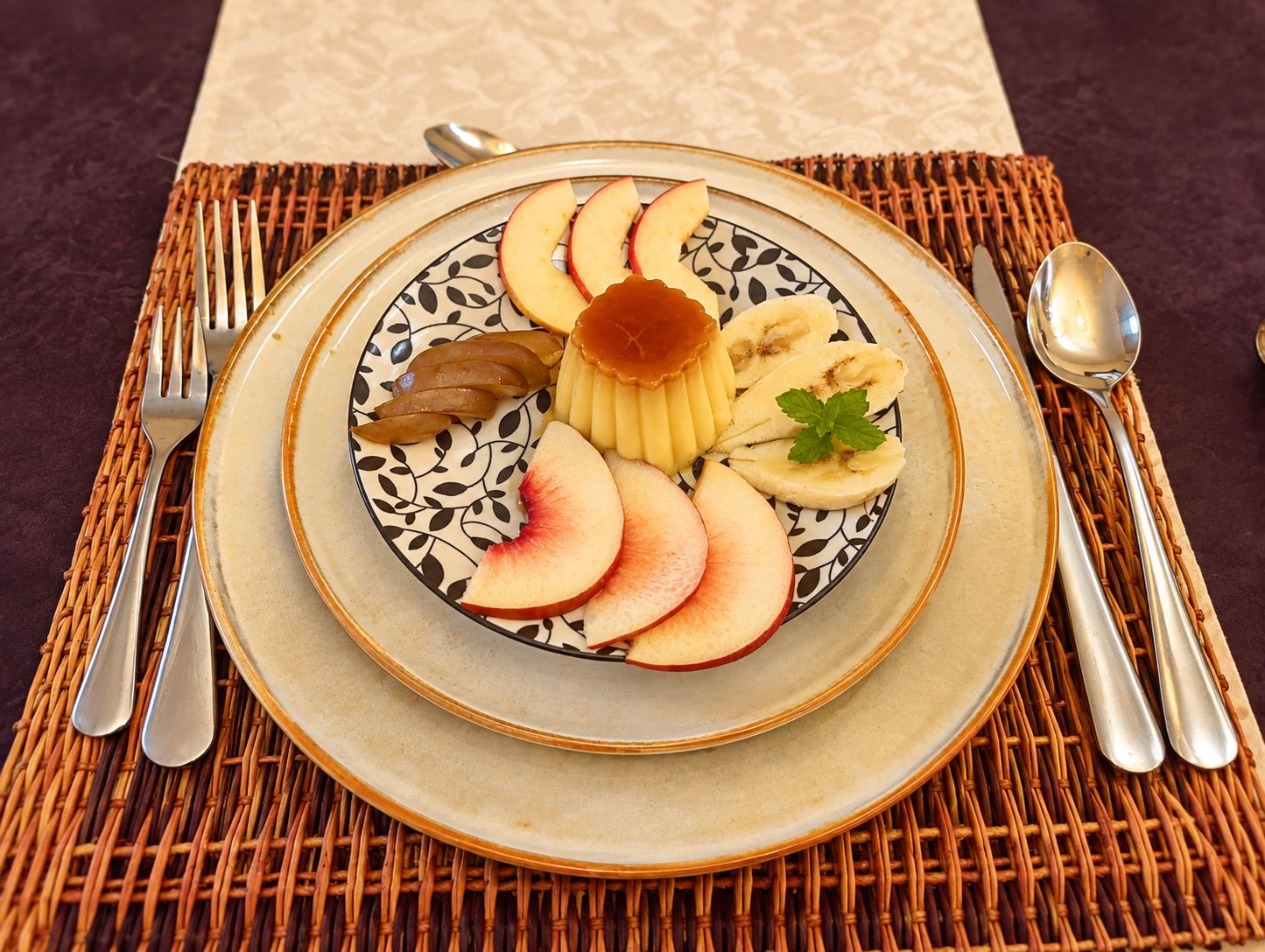 Un plato con rebanadas de manzanas, peras y plátanos, acompañado de una cuchara y un tenedor sobre un posavasos tejido.