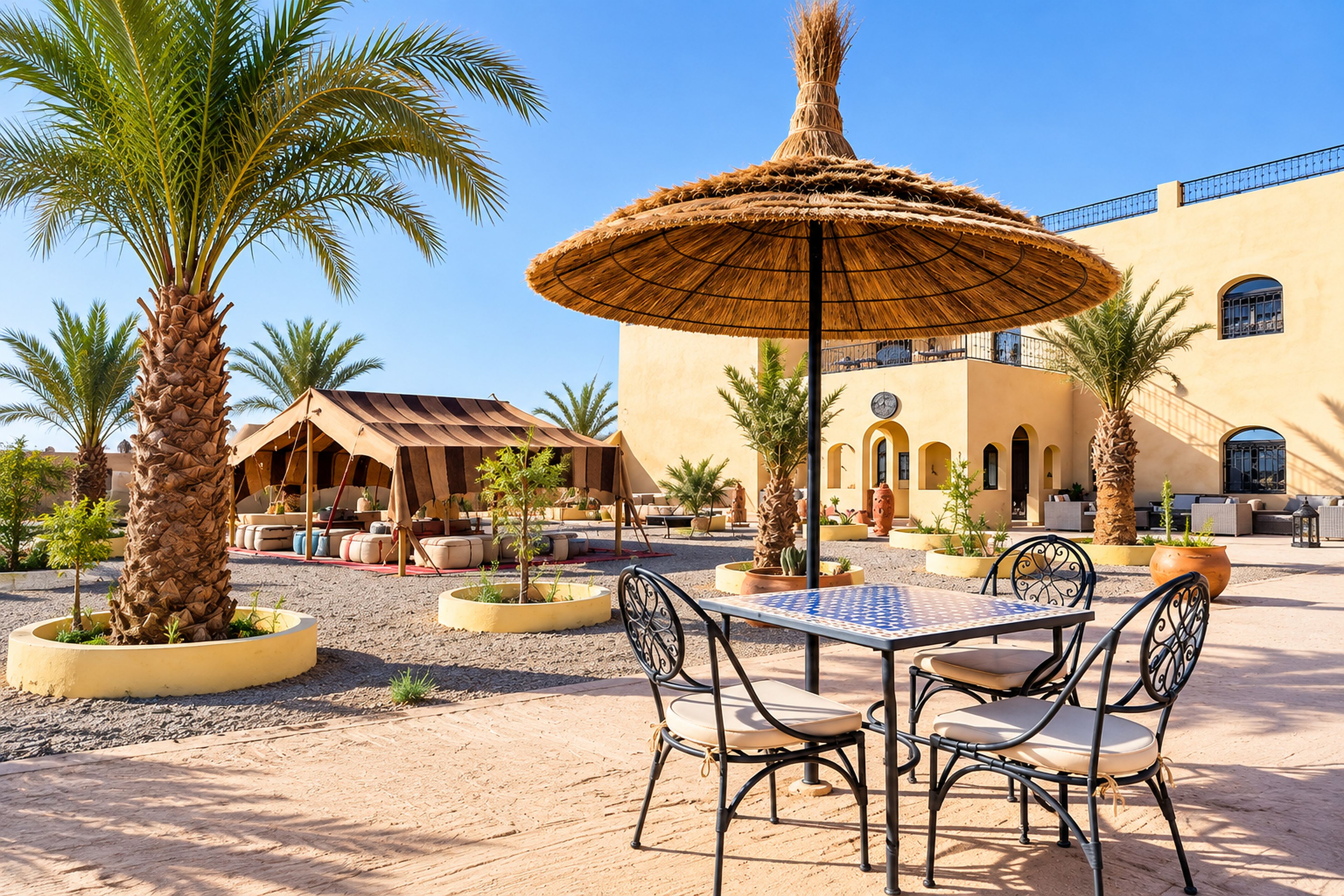 An outdoor seating area with a table and chairs, surrounded by palm trees and a building with arches.