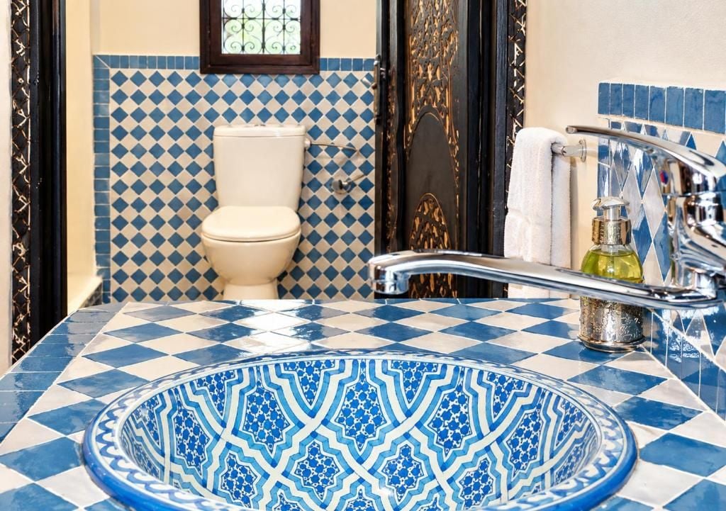 Un baño con un rebozado de azulejos blancos y azules a cuadros, un inodoro blanco, un lavamanos con una pileta de color azul estampada y una puerta dorada.