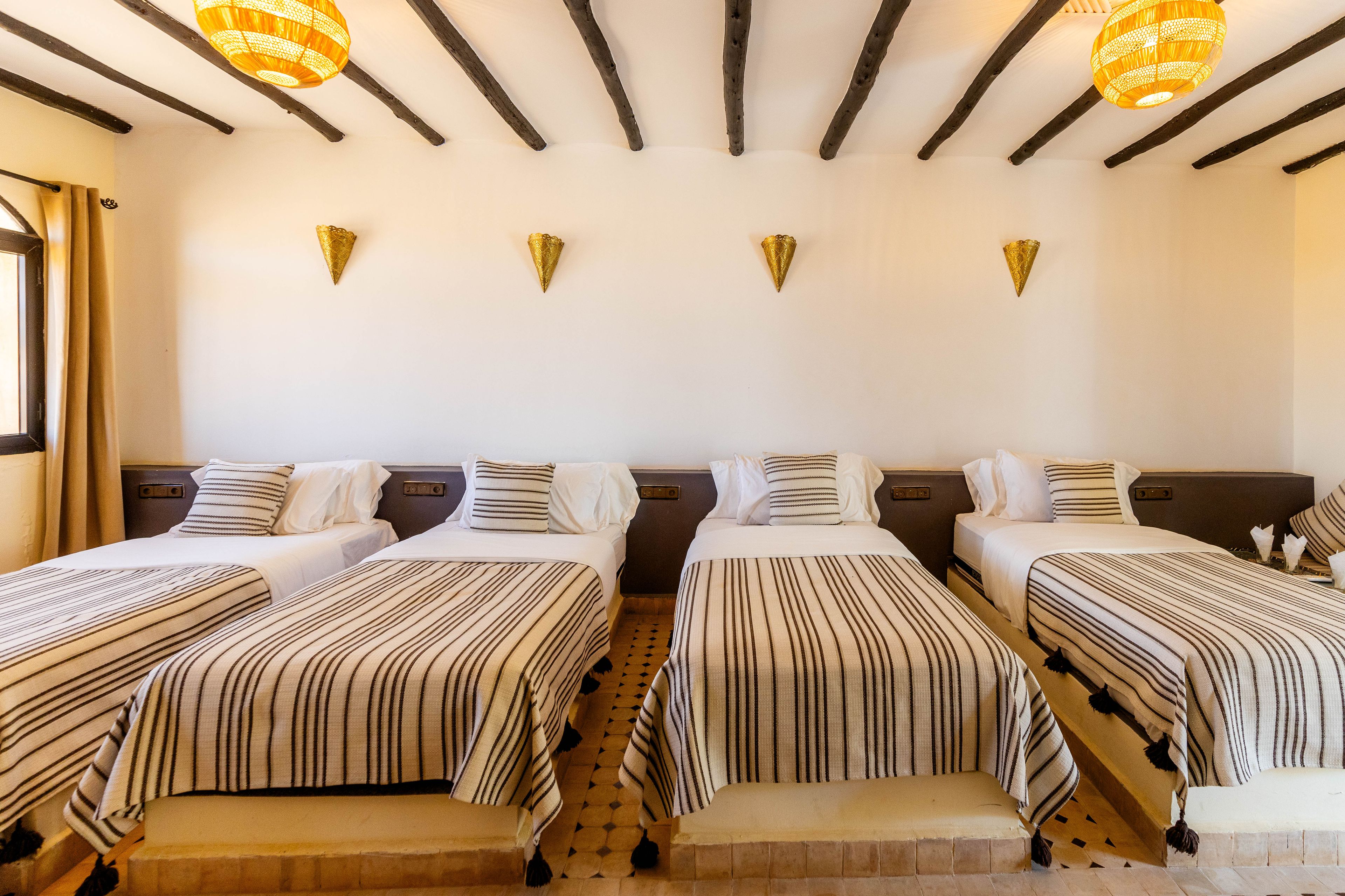Une chambre d’hôtel avec quatre lits simples, une grande fenêtre avec des rideaux et un plafond en bois avec des poutres apparentes.