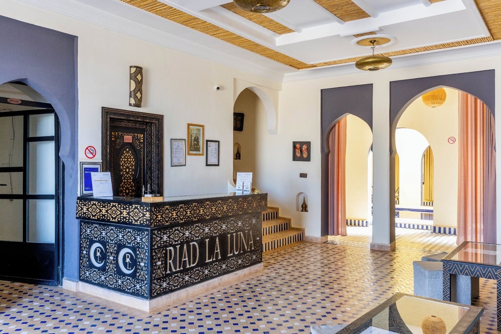 A reception area with a patterned floor, a reception desk with 'RIAD LA LUNA' signage, framed certificates on the wall, and a view of the courtyard through an archway.