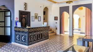 A reception area with a patterned floor, a reception desk with 'RIAD LA LUNA' signage, framed certificates on the wall, and a view of the courtyard through an archway. - Riad La Luna (Merzouga)
