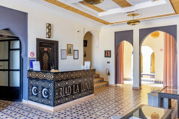 A reception area with a patterned floor, a reception desk with 'RIAD LA LUNA' signage, framed certificates on the wall, and a view of the courtyard through an archway. - Riad La Luna (Merzouga)