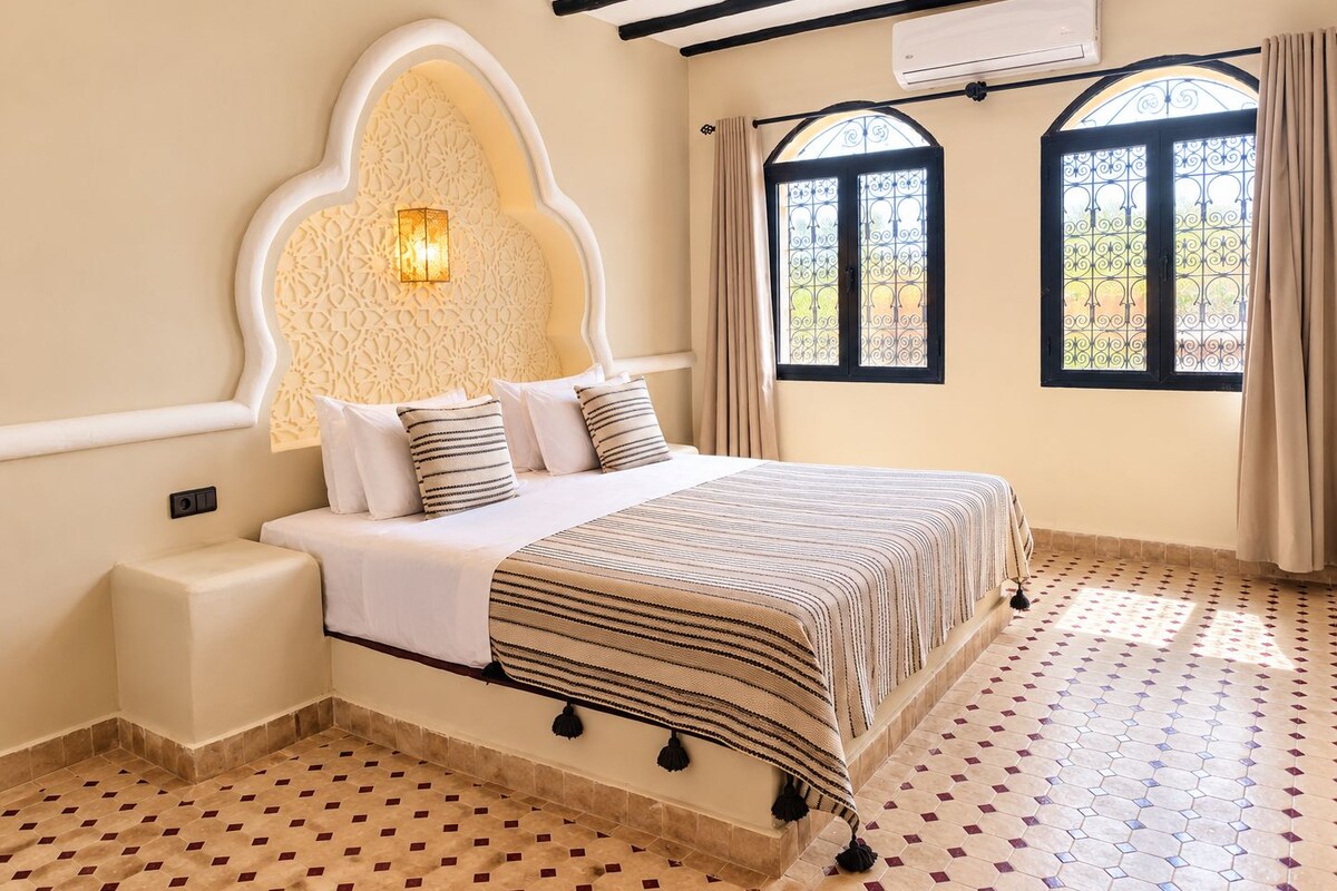 Quarto com cama, luminária fixada na parede, janela com grade de metal e piso estampado.