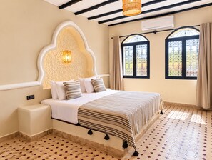 A room with a bed, a wall-mounted light, a window with a metal grill, and a patterned floor. - Riad La Luna (Merzouga)