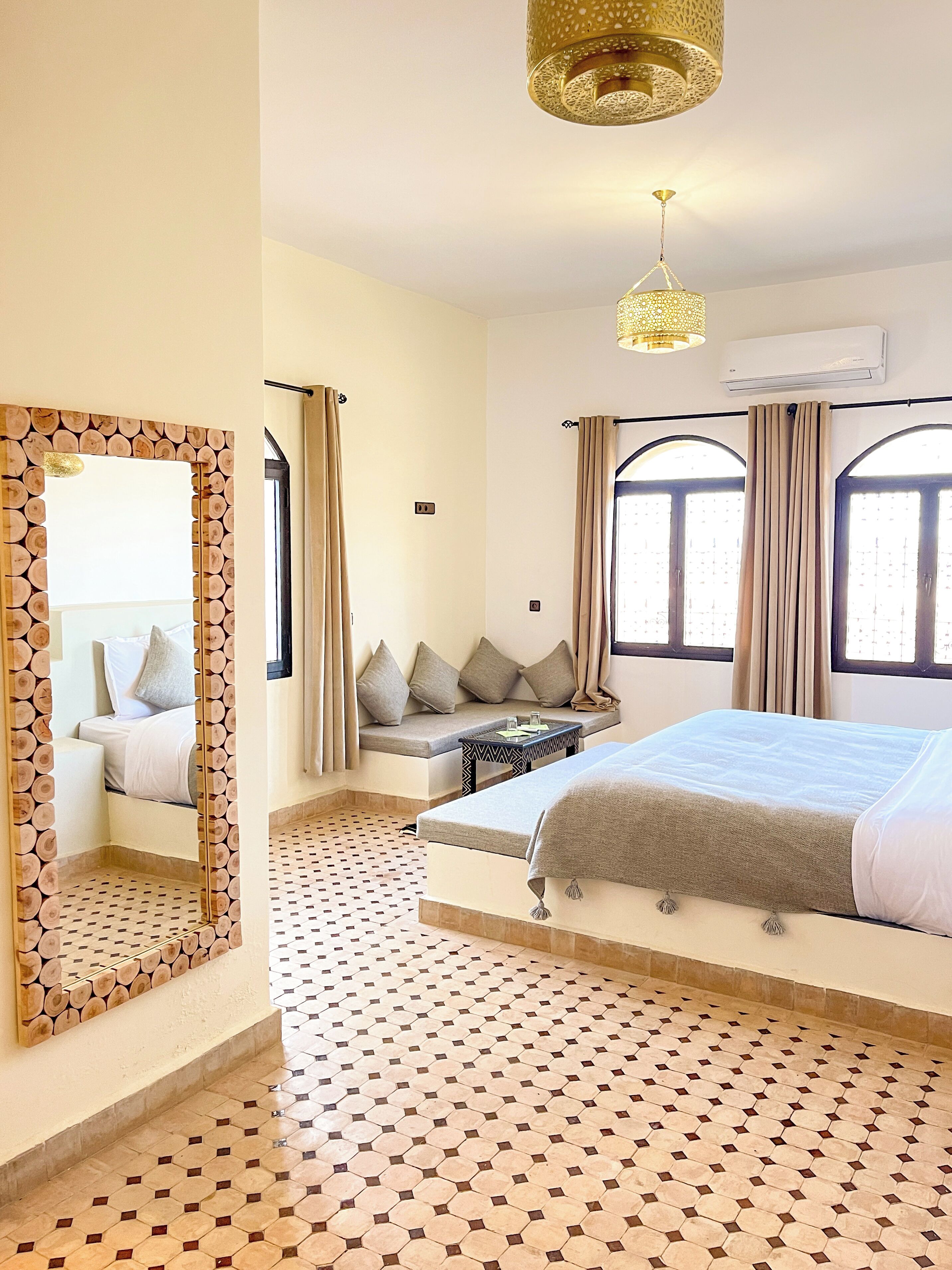 A hotel room with a large bed, a seating area with cushions, a patterned floor, and a large mirror with a wooden frame.