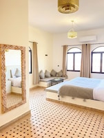 Una habitación de hotel con una cama grande, una zona de estar con cojines, un suelo estampado y un espejo grande con marco de madera.