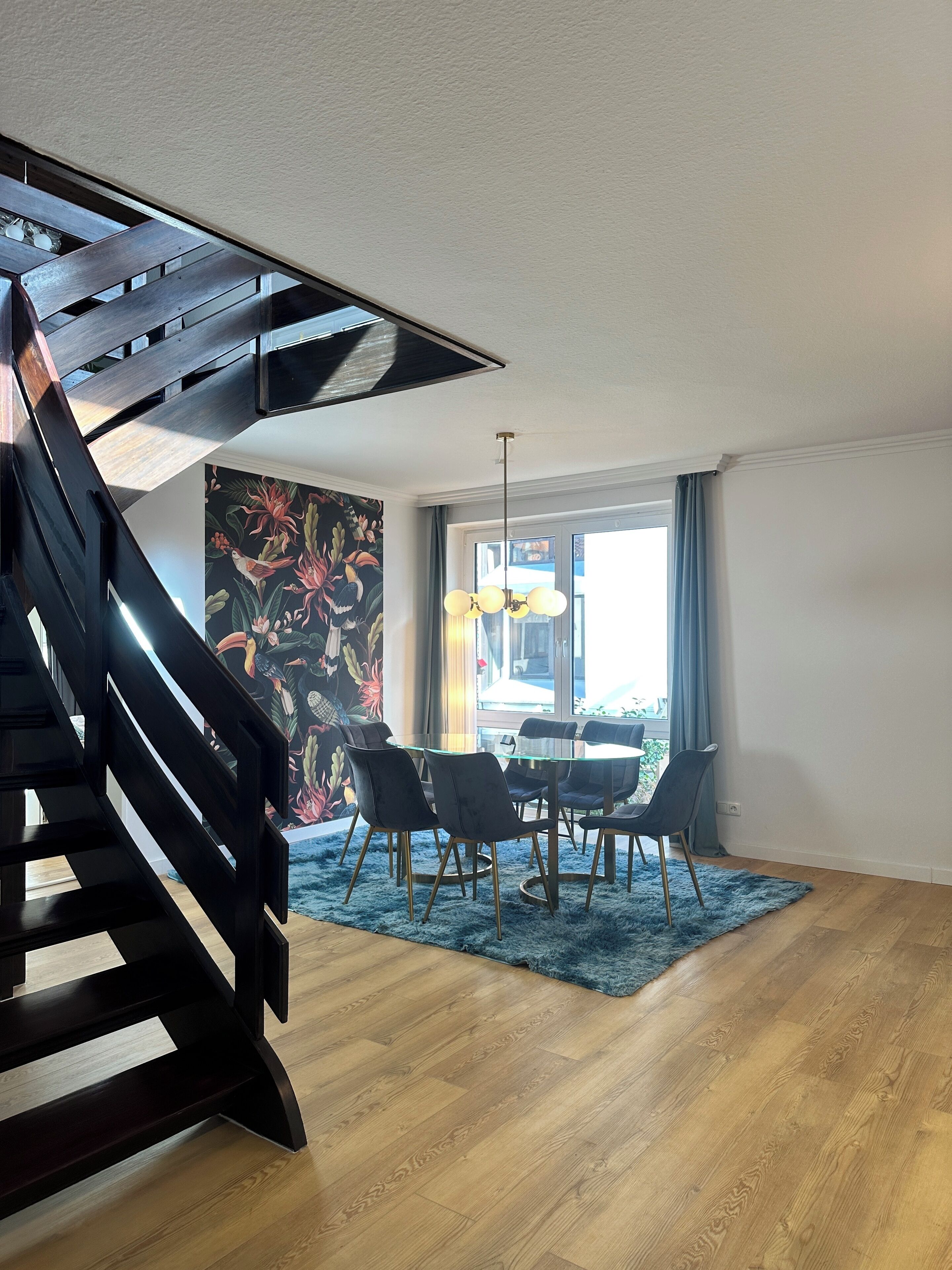 A modern interior with a wooden staircase, a dining area with a glass table and chairs, and a large window with curtains.