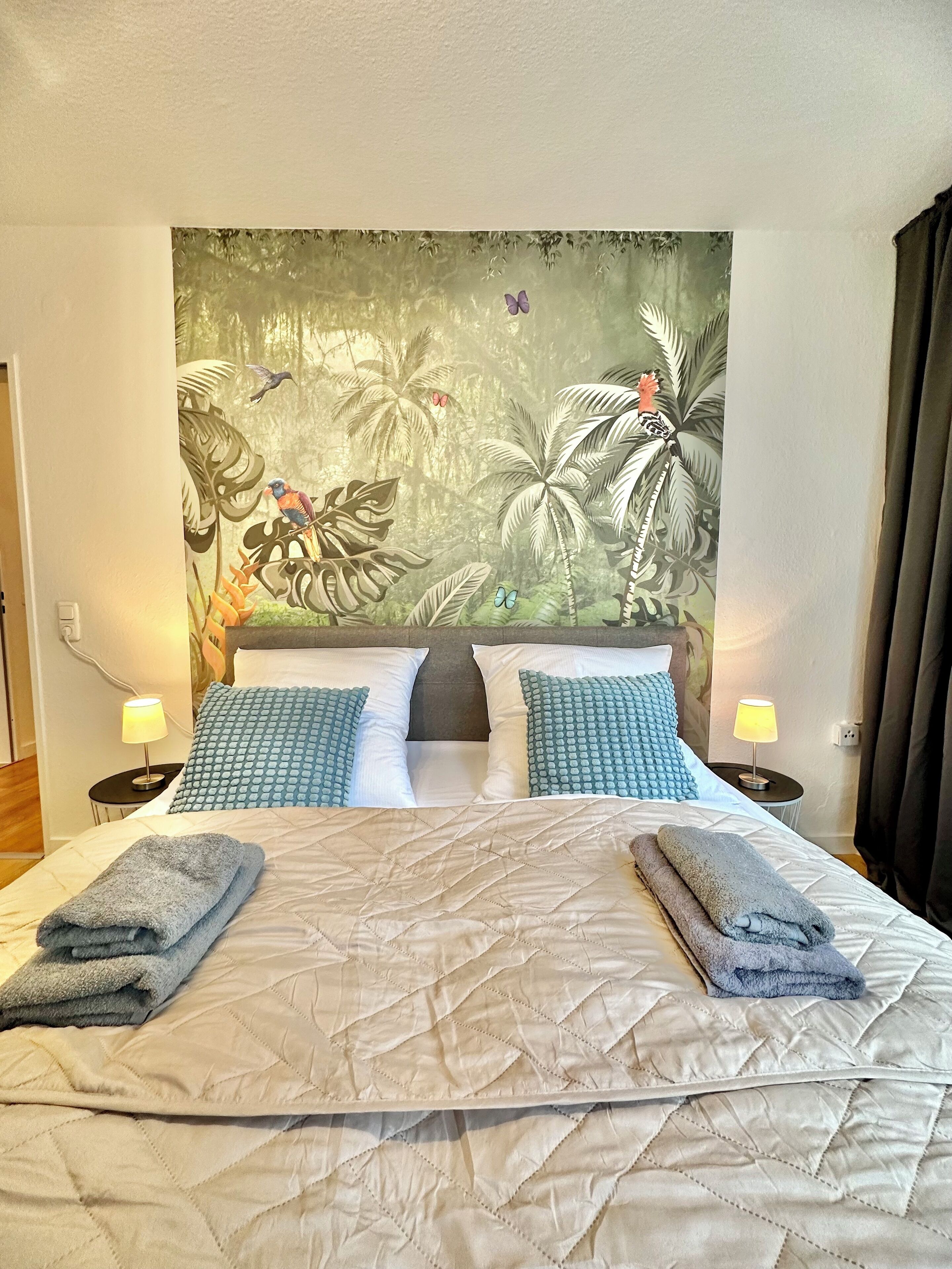 Un lit double avec une tête de lit matelassée, deux tables de chevet avec des lampes et une fresque murale sur le thème des tropiques.