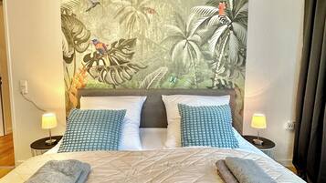 Un lit double avec une tête de lit matelassée, deux tables de chevet avec des lampes et une fresque murale sur le thème des tropiques.
