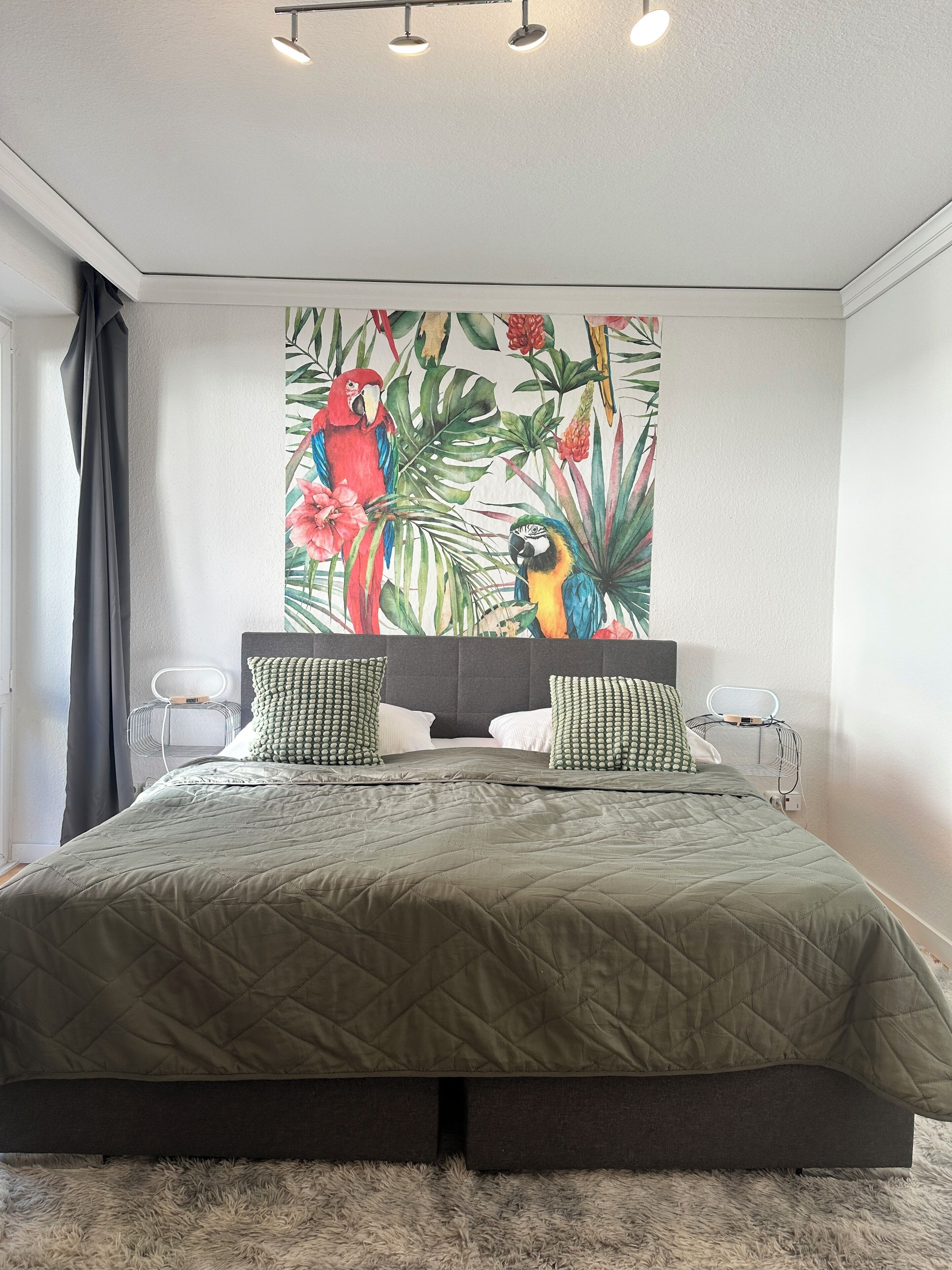 Ein Schlafzimmer mit einem großen Bett, zwei Kissen, einer Wandleuchte und einem Wandbild mit tropischem Motiv.