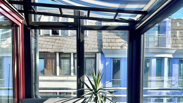 En modern utomhusplats med en soffa och ett litet bord, en krukväxt och utsikt över stadsbilden genom stora fönster.