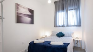 A bedroom with a bed, a bedside table with a lamp, a window with curtains, and a wall-mounted picture.