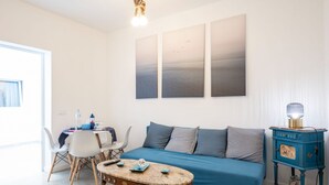 A modern living room with a blue sofa, a wooden coffee table, and a dining area with white chairs. There are three paintings on the wall, a lamp on a side table, and a window providing natural light.
