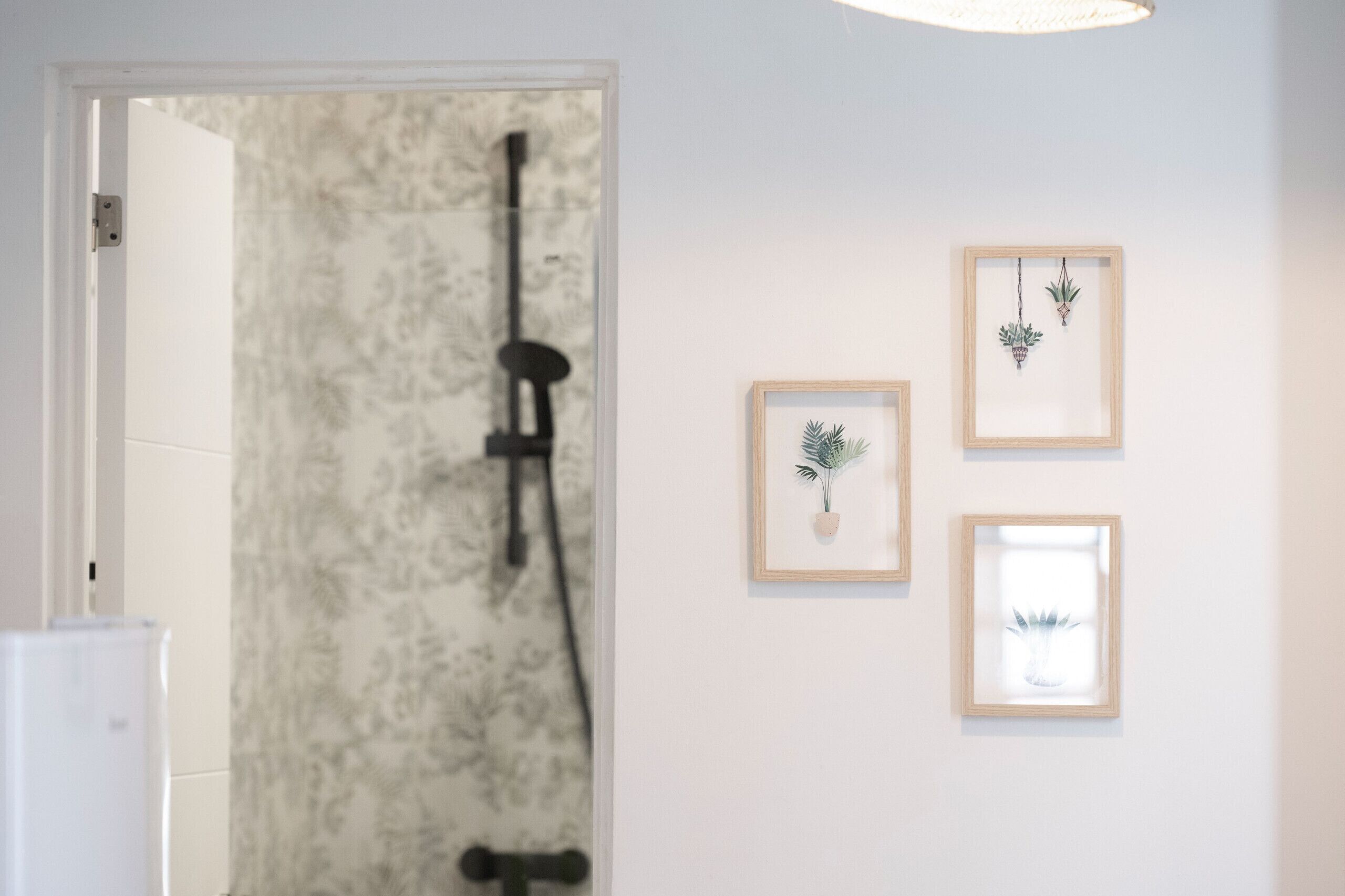 Et badeværelse med en bruseniche, en hvid dør og tre indrammede botaniske tryk på væggen.