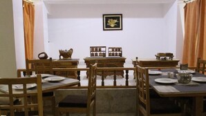 A dining area with wooden tables and chairs, a framed picture on the wall, and a window with curtains. - The Nun Kun Residency Kargil (Kargil)