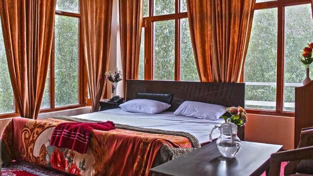 A bedroom with a large bed, a bedside table with a vase, and curtains on the windows. - The Nun Kun Residency Kargil (Kargil)