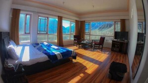 A hotel room with a large bed, wooden flooring, a desk with a chair, a television, and large windows offering a mountain view. - The Nun Kun Residency Kargil (Kargil)