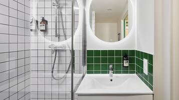 Une salle de bain avec des murs recouverts de carreaux blancs, un mur d’accent orné de carreaux verts, un lavabo blanc, une douchette à main et un miroir rond.