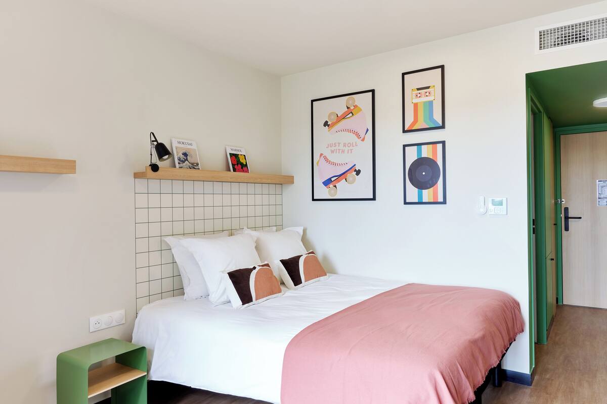 Een keurig opgemaakt slaapkamer met een bed, kussens, een plank met boeken en lijstjes met kunstwerken aan de muur.