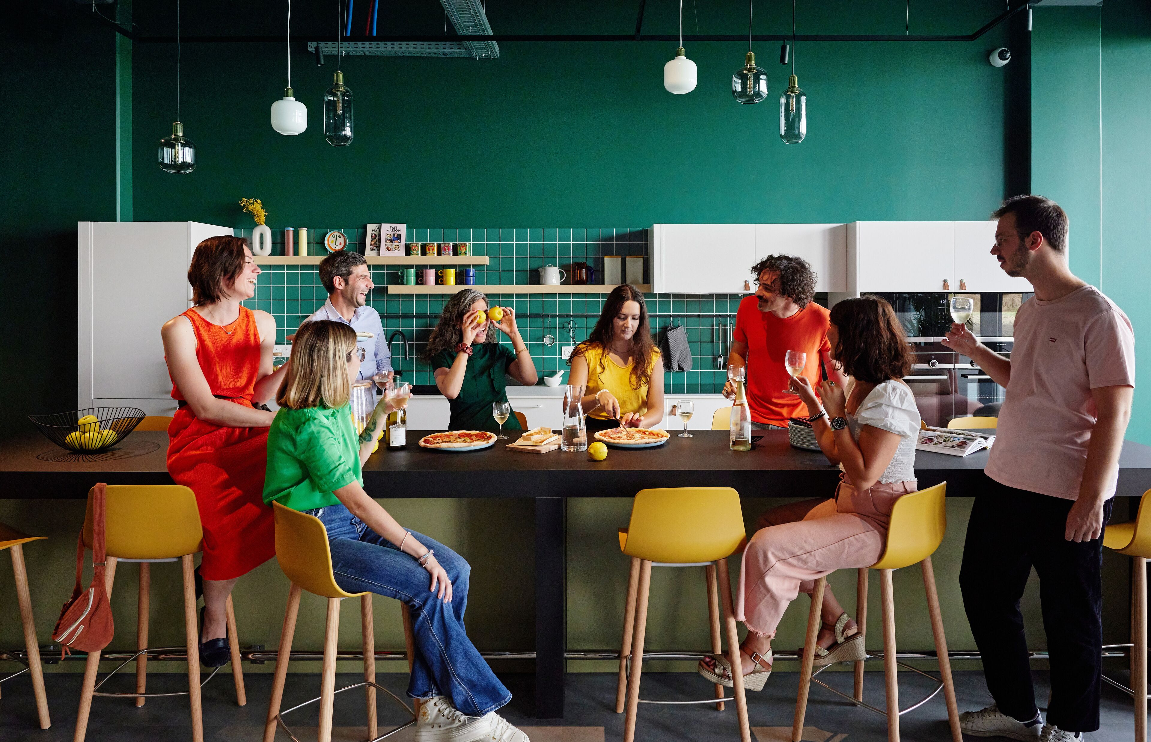 A group of people seated around a table in a modern kitchen with green walls and yellow chairs.