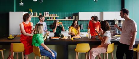 Un groupe de personnes est assis autour d’une table dans une cuisine moderne aux murs verts et aux chaises jaunes.