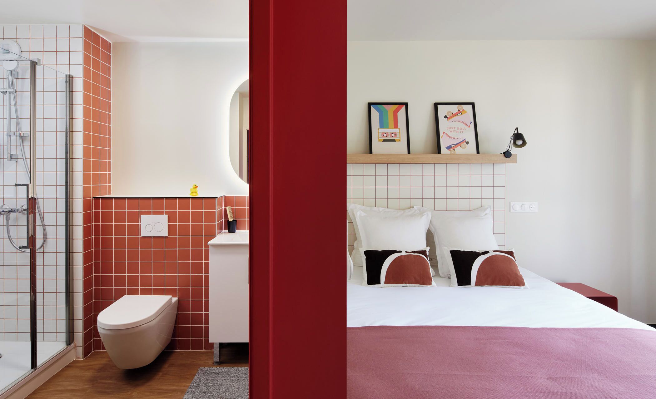 A bathroom with a shower, toilet, and sink. There is a bed with pillows and a bedside table.