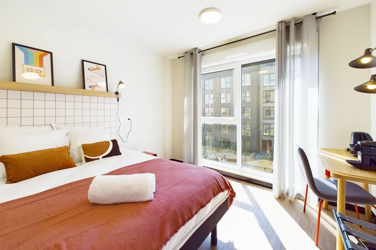 Een hotelkamer met een groot bed, een bureau met stoel, een raam met uitzicht op gebouwen en een plafondlamp.