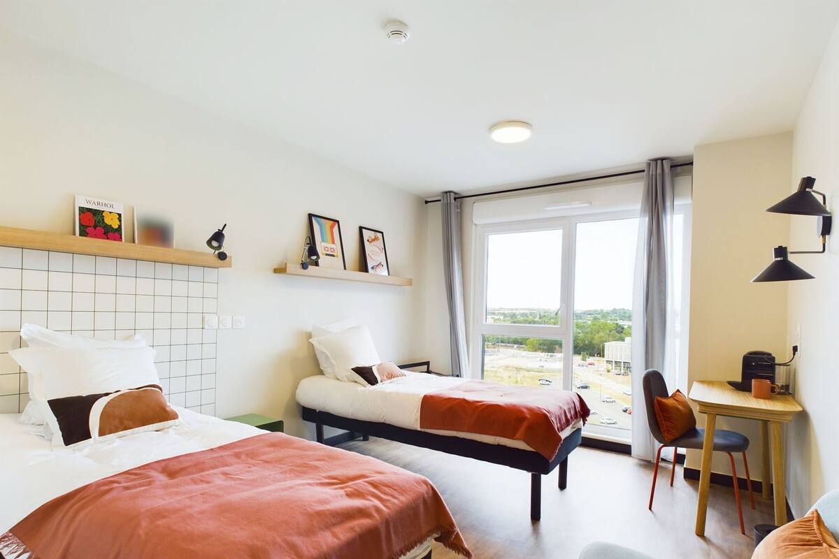 Hotelkamer met twee bedden, een bureau en een stoel. Er hangen prenten aan de muur, een raam met uitzicht en een plafondlamp.