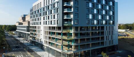 A modern multi-story building with a glass facade, multiple balconies, and a parking area in front.
