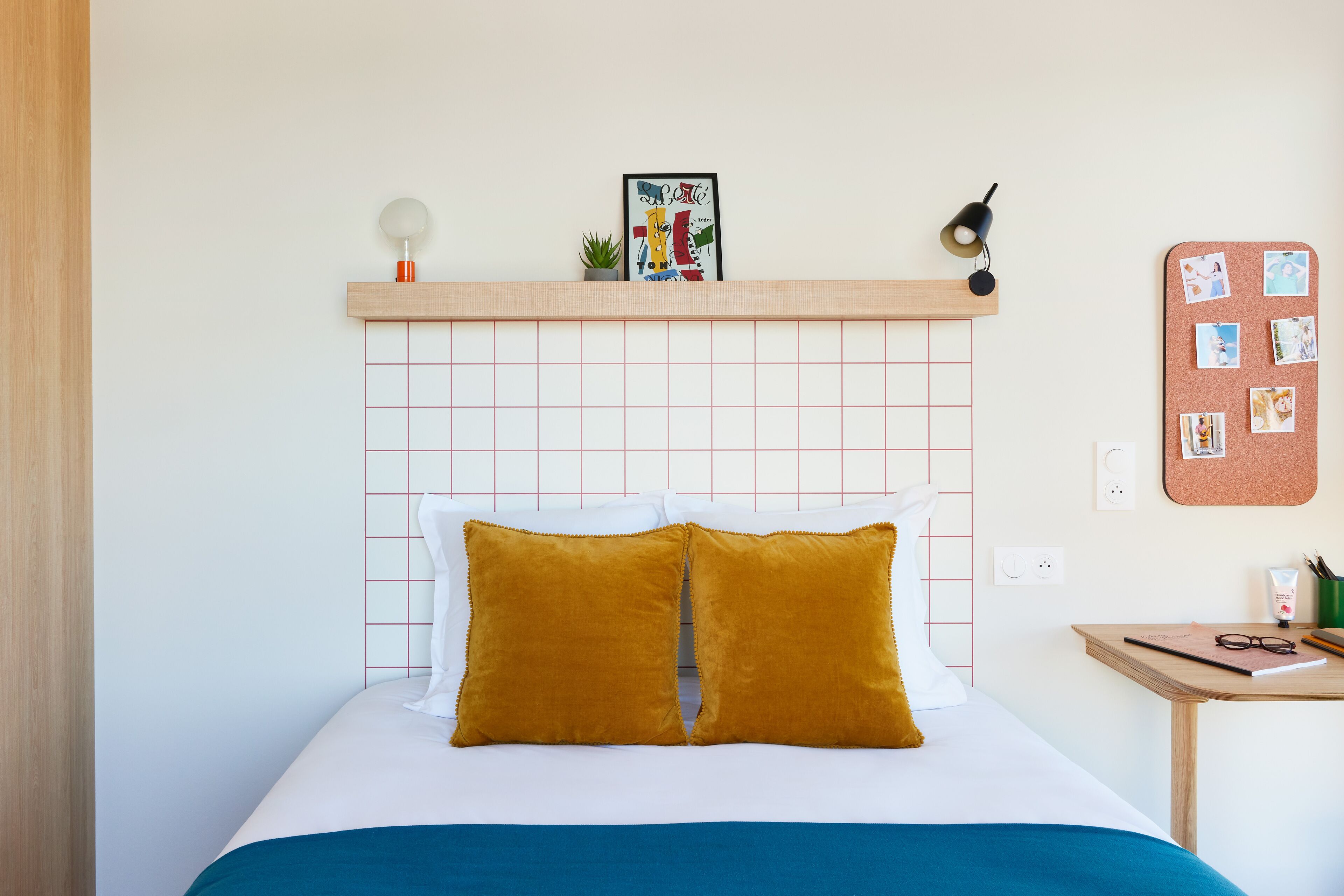 A neatly made bed with white and mustard pillows, a cork bulletin board with photos, and a small table with a lamp and a plant.