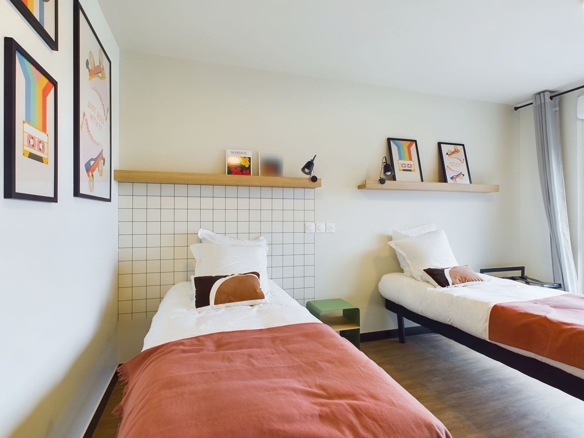 Una habitación de hotel con dos camas, cada una con ropa blanca y una manta marrón. Hay cuadros enmarcados en la pared y un estante con un grabado de Warhol y una lámpara.