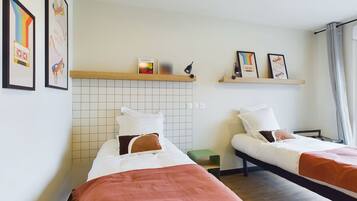 Una habitación de hotel con dos camas, cada una con ropa blanca y una manta marrón. Hay cuadros enmarcados en la pared y un estante con un grabado de Warhol y una lámpara.