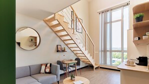 Un espacio habitable moderno con una escalera, un espejo redondo, un sofá gris, una mesita con una planta y vistas a través de grandes ventanales.