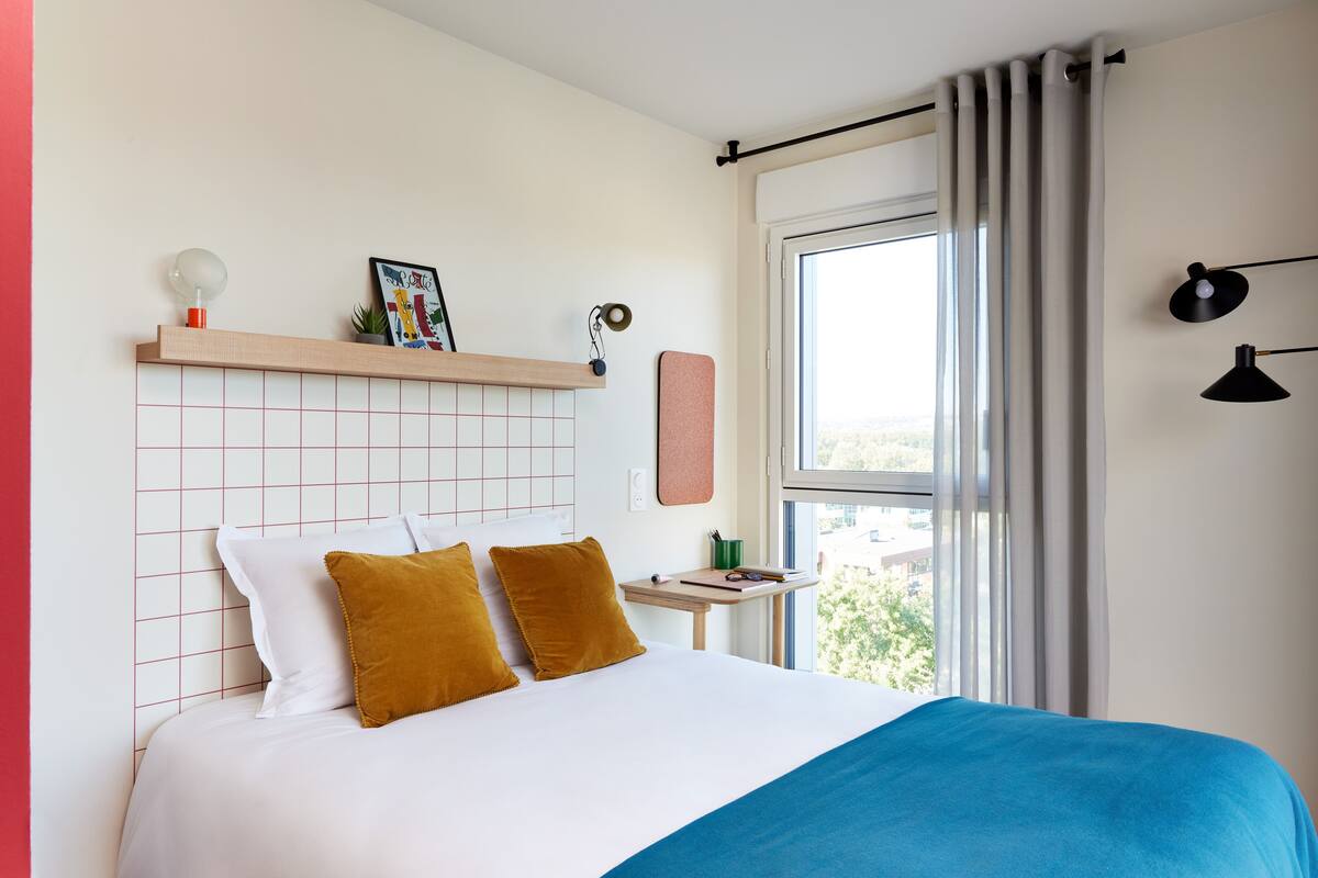 Een netjes opgemaakt bed met wit en blauw beddengoed, een klein nachtkastje met een lamp en een raam met gordijnen.