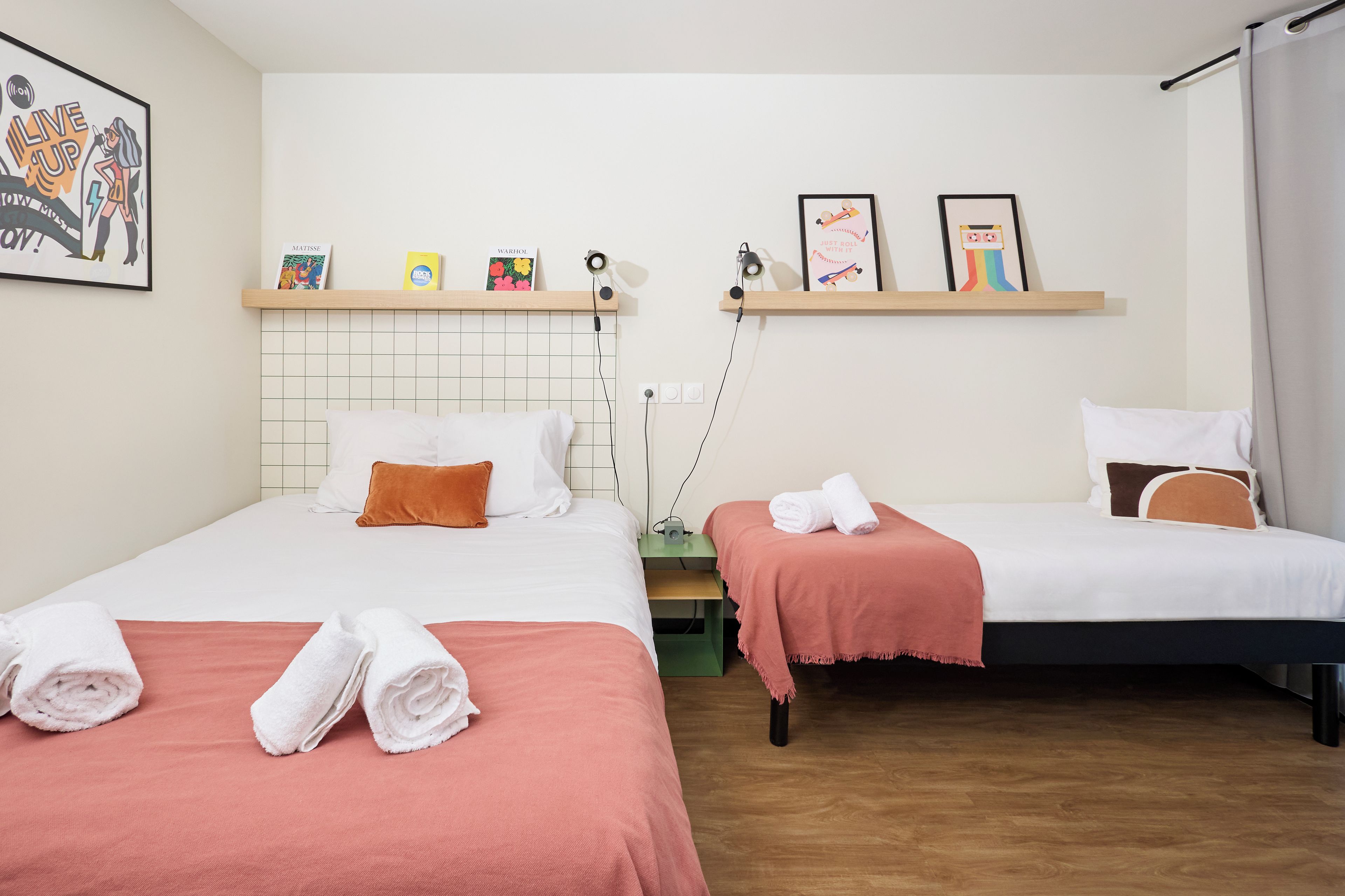 Habitación de hotel con dos camas, cada una con colcha blanca y rosa, cabecero de madera y repisa sobre las camas con libros y una lámpara.