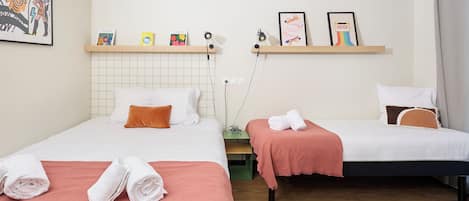 Habitación de hotel con dos camas, cada una con colcha blanca y rosa, cabecero de madera y repisa sobre las camas con libros y una lámpara.