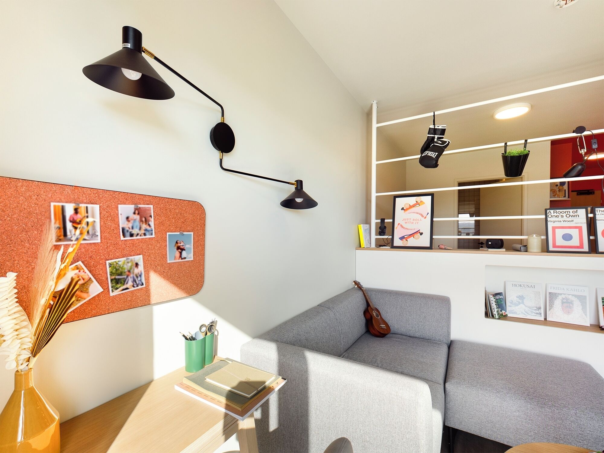 Une pièce chaleureuse avec un canapé gris, un tableau d’affichage en liège où sont accrochées des photos, une petite table avec un livre et un pot vert, et une étagère murale où sont posés des livres et un tableau encadré.