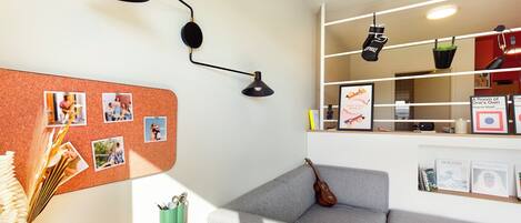Une pièce chaleureuse avec un canapé gris, un tableau d’affichage en liège où sont accrochées des photos, une petite table avec un livre et un pot vert, et une étagère murale où sont posés des livres et un tableau encadré.